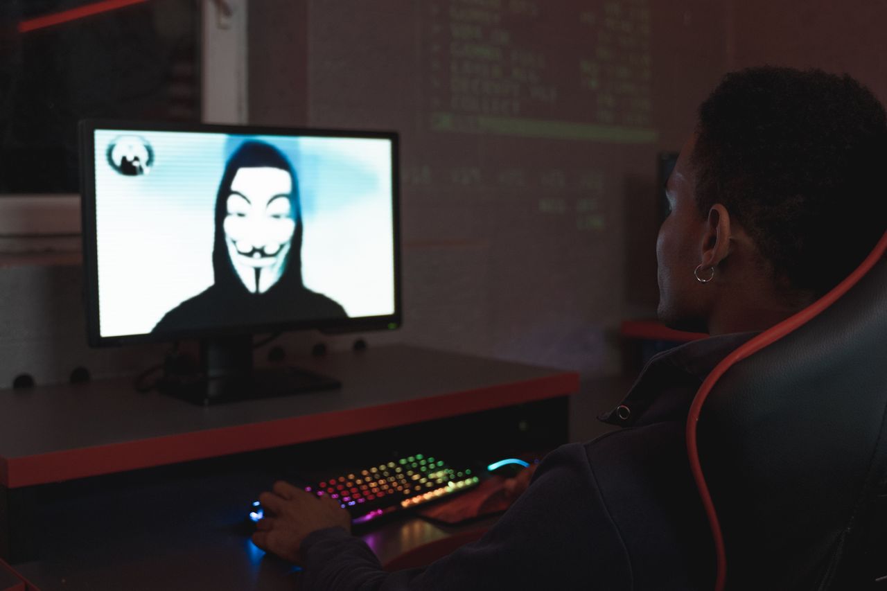 Person using computer in dark room viewing hacker wearing a mask on the screen, symbolizing cyber security threats.